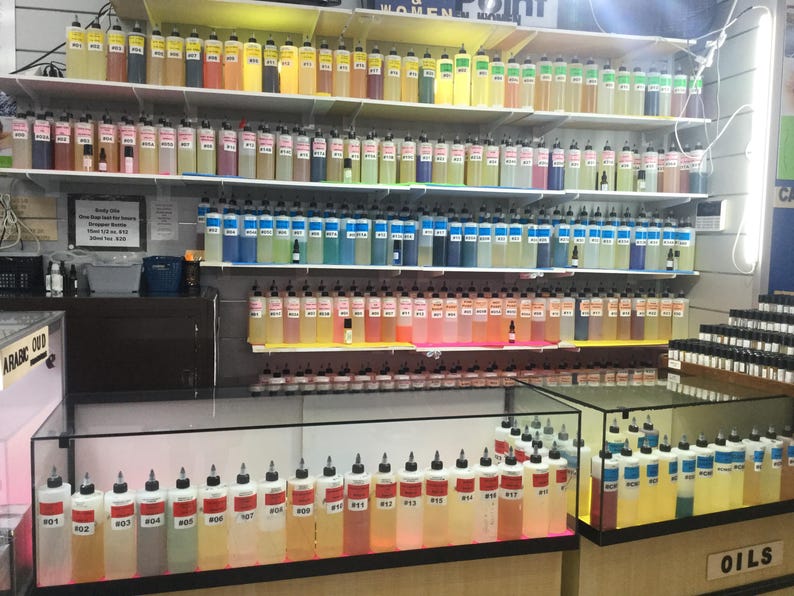 May include: A display of various colorful oils in clear bottles with black caps, arranged on shelves and in glass display cases. The bottles are labeled with numbers and contain liquids in shades of yellow, pink, blue, and more. The word "OILS" is visible.