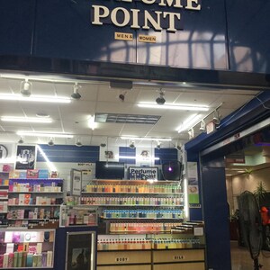 May include: The entrance to a perfume store, "Perfume Point," is visible, with the store's name in large white letters above the entrance. The store sells body oils and perfumes for men and women. Shelves are filled with various perfume bottles.