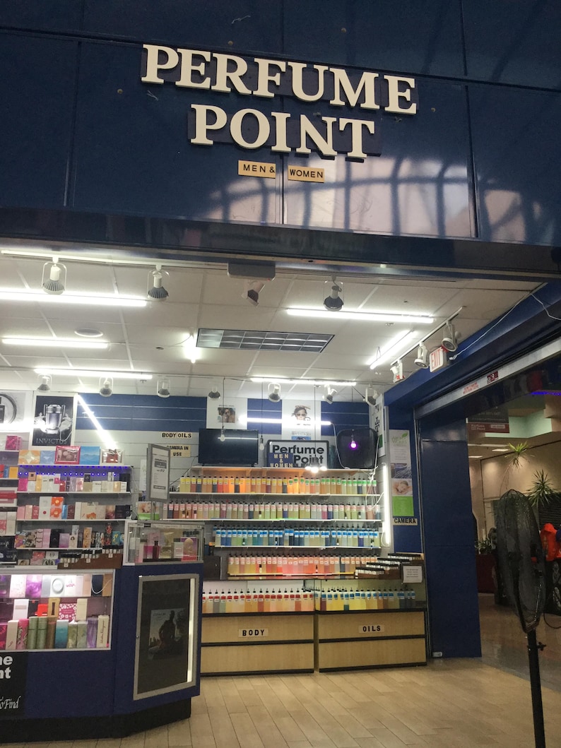 May include: The entrance to a perfume store, "Perfume Point," is visible, with the store's name in large white letters above the entrance. The store sells body oils and perfumes for men and women. Shelves are filled with various perfume bottles.