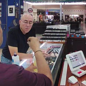 May include: Two men are in a store, one is holding a phone and the other is holding a bottle. A display case with many small bottles is behind them. A small computer screen is on a table.
