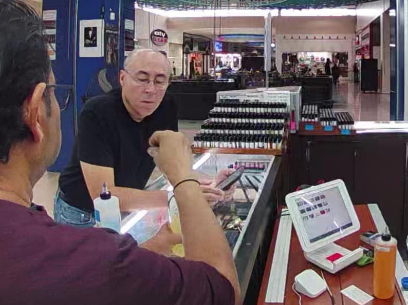 May include: Two men are in a store, one is holding a phone and the other is holding a bottle. A display case with many small bottles is behind them. A small computer screen is on a table.