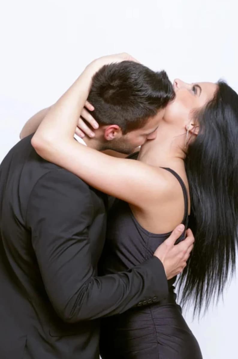 May include: A couple in an embrace, the man in a black suit and the woman in a black dress. The man is kissing the woman's neck, and they are close together. The woman has long, dark hair.