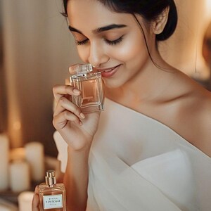 May include: A woman holding a clear perfume bottle with a rectangular shape and a clear cap. The bottle is held up to her nose. Another perfume bottle is visible in the foreground. The woman is wearing a white dress.