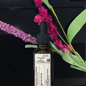 May include: Aroma Works perfume bottle with a black dropper, a white label, and the text "EXTRAIT DE PARFUM". The bottle is brown, and the label includes the text "One dap last for hours". The background includes pink and purple flowers.
