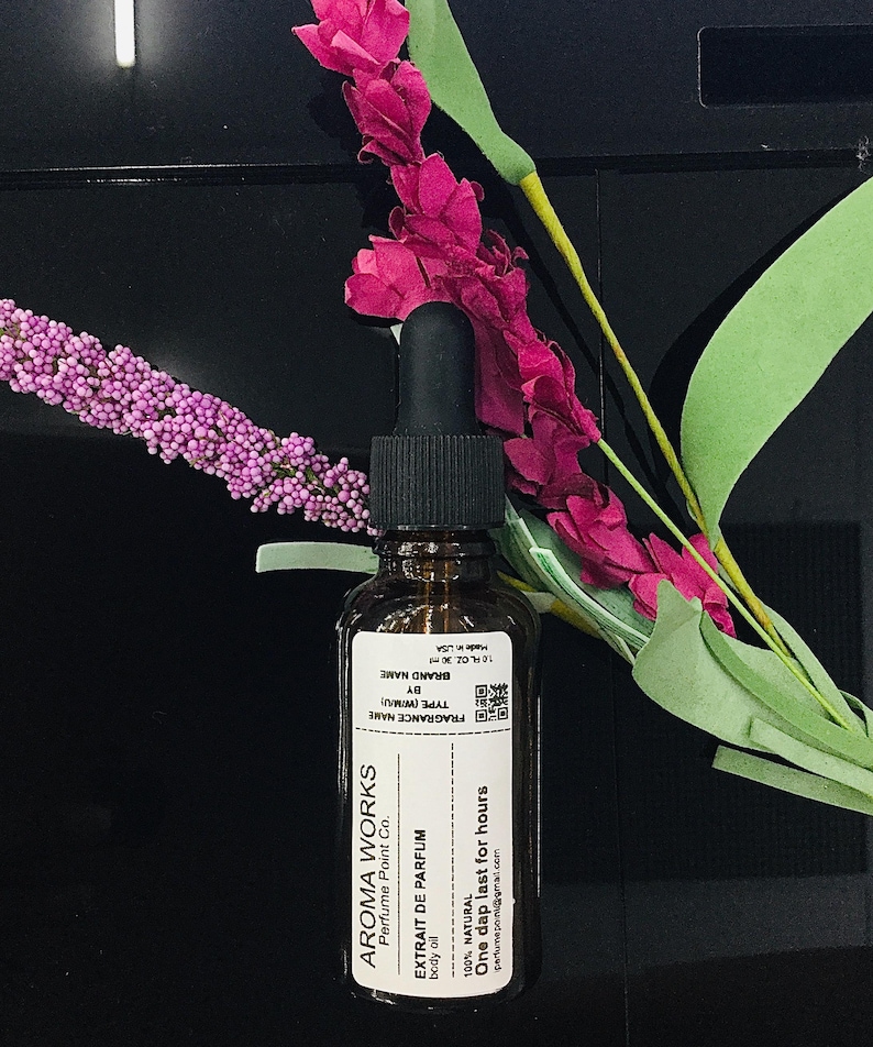 May include: Aroma Works perfume bottle with a black dropper, a white label, and the text "EXTRAIT DE PARFUM". The bottle is brown, and the label includes the text "One dap last for hours". The background includes pink and purple flowers.