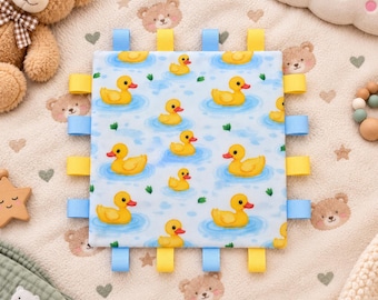 Könnte beinhalten: Eine quadratische Taggie-Babydecke mit einem wiederkehrenden Muster aus gelben Quietscheentchen auf hellblauem Wasserhintergrund. Die Decke hat mehrere gelbe und hellblaue Bandschlaufen an allen vier Seiten für taktiles sensorisches Spielen.