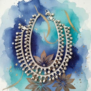May include: A pair of silver-toned anklets featuring a flat, woven chain design adorned with numerous small, dangling spherical bells. Each anklet includes a secure lobster claw clasp and a small circular charm attached near the closure mechanism.