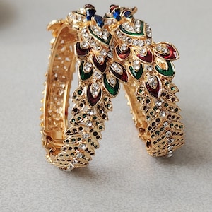 May include: Two gold-colored bangles with intricate peacock feather designs. The bangles are adorned with red, green, blue, and clear gemstones. The peacock feather design is at the top of the bangles.