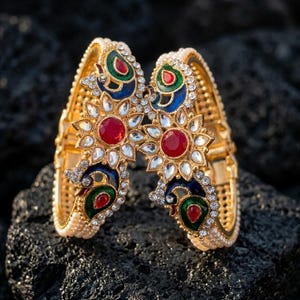 Könnte beinhalten: Zwei goldene Armreifen mit aufwendigen Pfau- und Blumenmustern. Jeder Armreif hat einen zentralen roten Edelstein, umgeben von klaren Kristallen und Emaille-Details in Blau, Grün und Rot. Auf einem dunklen, strukturierten Hintergrund.
