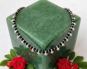 May include: A delicate necklace featuring black beads and silver-colored accents. The necklace is displayed on a green, square-shaped object, with red flowers and green leaves in the foreground. The necklace is likely made of metal and gemstones.