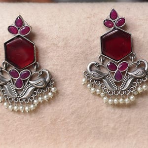 May include: A pair of oxidized silver earrings featuring a central hexagonal deep red stone, a smaller three-petal red stone cluster, and a base adorned with stylized bird motifs and a fringe of small white beads hanging from the bottom edge.