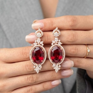 May include: Rose gold drop earrings featuring large oval-cut red gemstones surrounded by a halo of small, clear crystals. Additional clear crystals adorn the top and bottom of the earrings, creating a sparkling, elegant design.
