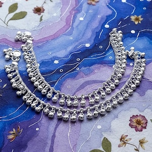 May include: Two silver anklets with small bells and intricate chain detailing. The anklets are displayed on a blue and purple watercolor background with dried flowers, creating a delicate and bohemian aesthetic.