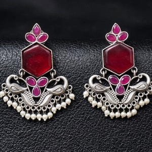 May include: A pair of silver-toned earrings featuring a central hexagonal red stone, a smaller three-petal red stone cluster, and two stylized bird motifs. The lower edge is decorated with a row of small white beads hanging from the metal frame.