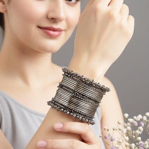 Può includere: Una pila di braccialetti color argento con bordi perlati adorna un polso. I braccialetti sono indossati su un braccio, con uno sfondo grigio e un bouquet di fiori bianchi e viola nell'angolo.