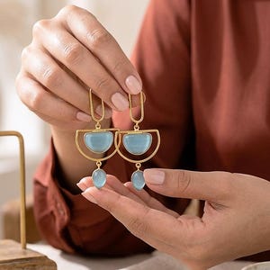 Könnte beinhalten: Goldfarbene Ohrringe mit einem Halbmond-Design und einem hellblauen, oval geformten Stein. Die Ohrringe werden von einer Person hochgehalten. Die Ohrringe haben einen modernen, geometrischen Stil.