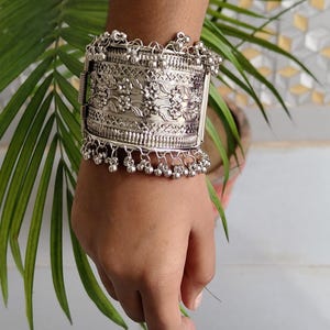 Könnte beinhalten: Silberfarbenes Armband mit aufwendigen floralen und geometrischen Mustern. Das Armband hat baumelnde Glockenanhänger und einen Klappverschluss. Das Design ist detailliert und verziert, geeignet als auffälliges Schmuckstück.