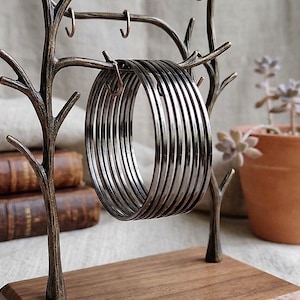 Può includere: Un espositore per gioielli color bronzo a forma di albero, che tiene una pila di braccialetti d'argento. L'espositore ha una base in legno e ganci per appendere i gioielli. Una piccola pianta in vaso è visibile sullo sfondo.
