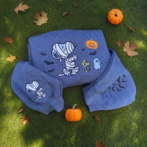 Pode incluir: Um moletom azul com um design de Snoopy múmia, uma abóbora pequena e um fantasma. A manga tem um design de morcego. O moletom está na grama verde, com folhas e abóboras pequenas.