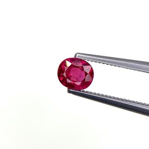 May include: A vibrant, oval-cut ruby gemstone held by silver tweezers. The ruby displays a deep red hue and a faceted surface, reflecting light. The gemstone is set against a white background.