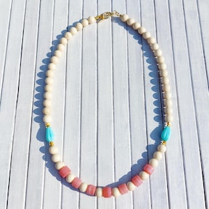 Peut inclure: Collier de perles crème avec des accents de goutte d'eau turquoise et des perles carrées roses. Le collier a un fermoir doré et est présenté sur une surface en bois blanc.