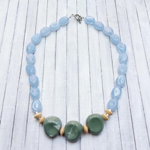 Op de afbeelding: Een ketting met lichtblauwe ovale kralen en drie grotere, olijfgroene kralen. Kleine, lichtbruine kralen scheiden de groene kralen van de blauwe. De ketting heeft een zilverkleurige sluiting.