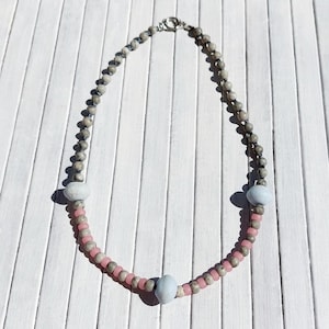 Op de afbeelding: Een ketting met een mix van grijze, roze en lichtblauwe kralen. De ketting heeft een zilveren sluiting en is samengesteld uit kleine, ronde grijze kralen, afgewisseld met grotere lichtblauwe en roze kralen.