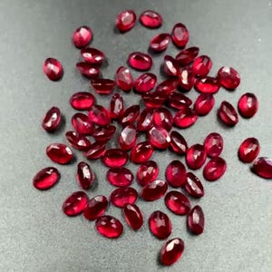 May include: A collection of oval-cut, deep red gemstones. The faceted stones are scattered across a gray surface, catching the light and displaying a rich, vibrant color. These gemstones are suitable for jewelry making.