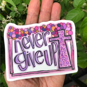 May include: A pink and purple sticker with the words "never give up" and a cross in the background. The cross is made up of small purple and pink dots.