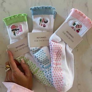 Könnte beinhalten: Drei Paar weiße Socken mit farbenfrohen Akzenten und Designs. Jedes Paar hat eine andersfarbige Manschette und ein einzigartiges gesticktes Motiv. Die Socken tragen den Text "REFORMED. FOR THE BETTER" auf den angebrachten Etiketten.