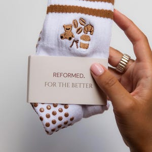 Könnte beinhalten: Weiße Socken mit braunen Streifen und Kaffee-Stickereien, darunter ein Bär, Kaffeebohnen und eine Tasse. Die Socken haben braune Anti-Rutsch-Punkte auf den Sohlen und ein Etikett mit der Aufschrift "REFORMED. FOR THE BETTER."