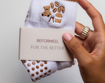 Pode incluir: Meias brancas com riscas castanhas e bordados com tema de café, incluindo um urso, grãos de café e uma chávena. As meias têm pontos antiderrapantes castanhos nas solas e uma etiqueta que diz "REFORMED. FOR THE BETTER."