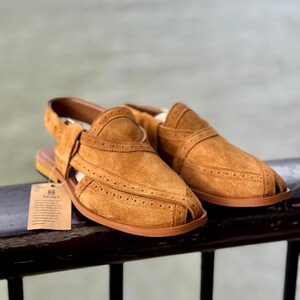 May include: A pair of tan suede sandals with a strap and open toe design. The sandals have decorative stitching and a brown sole. A small tag is attached to the left sandal.