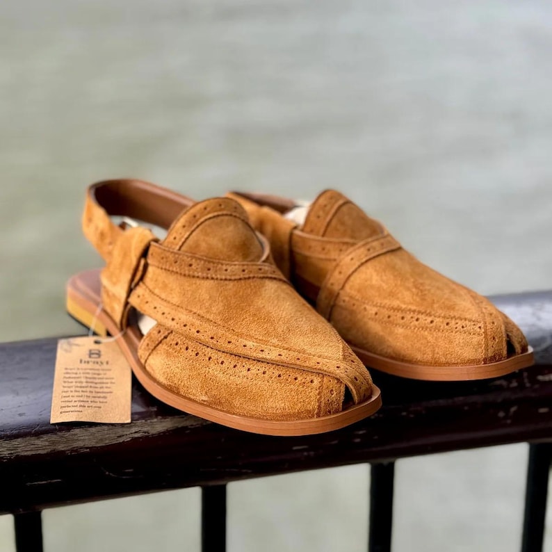 May include: A pair of tan suede sandals with a strap and open toe design. The sandals have decorative stitching and a brown sole. A small tag is attached to the left sandal.