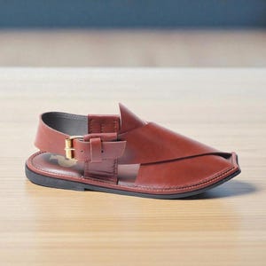 May include: Brown leather sandal with a buckle closure and a black sole. The sandal features a unique design with a strap across the top and a back strap for support. The footwear is designed for comfort and style.