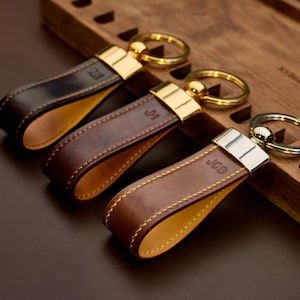 Puede incluir: Tres llaveros de cuero en tonos marrones, cada uno con un anillo de metal dorado o plateado. Los llaveros tienen iniciales personalizadas y costuras en contraste. Los llaveros se exhiben sobre una superficie de madera.