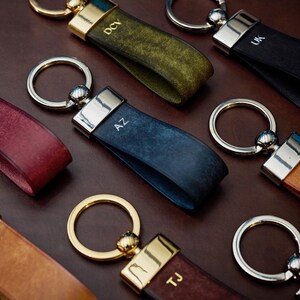 May include: A collection of leather keychains in various colors, including burgundy, olive green, navy blue, and tan. Each keychain features a metal ring and a strap with initials engraved on the leather. The keychains have silver and gold accents.