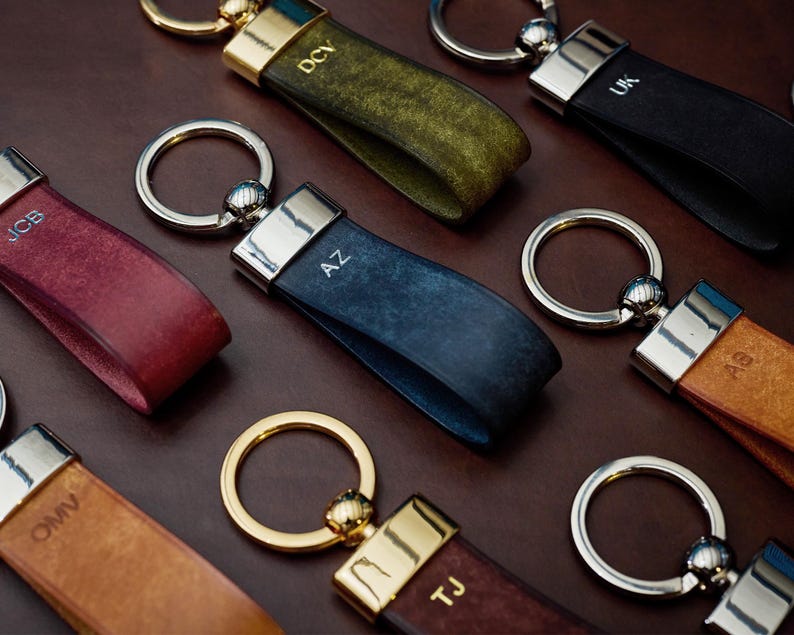 May include: A collection of leather keychains in various colors, including burgundy, olive green, navy blue, and tan. Each keychain features a metal ring and a strap with initials engraved on the leather. The keychains have silver and gold accents.