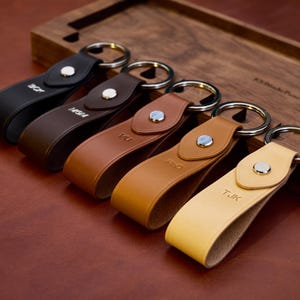 Peut inclure: Cinq porte-clés en cuir de couleurs noir, marron et beige, chacun avec un anneau porte-clés argenté et une fermeture à bouton-pression. Les porte-clés sont gravés d'initiales ou de chiffres. Ils sont disposés sur un plateau en bois.