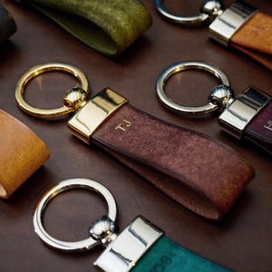 May include: Close-up of several leather keychains in various colors, including brown, teal, and burgundy. Each keychain features a metal ring and a leather strap with initials. The keychains are arranged on a dark brown surface.