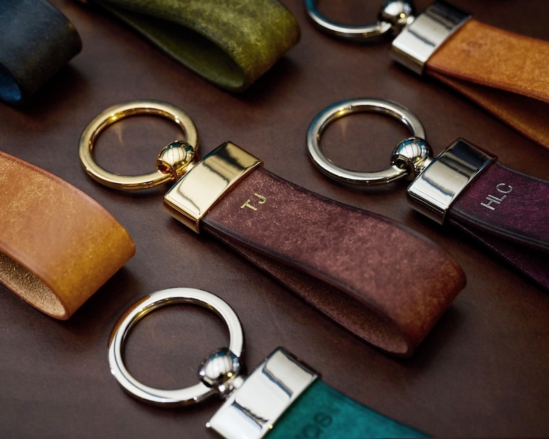 May include: Close-up of several leather keychains in various colors, including brown, teal, and burgundy. Each keychain features a metal ring and a leather strap with initials. The keychains are arranged on a dark brown surface.
