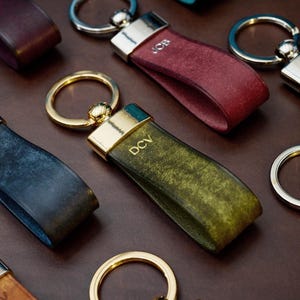 May include: Close-up of several leather keychains in various colors, including burgundy, green, and blue. Each keychain has a metal ring and a leather strap with initials engraved on the strap. The keychains are arranged on a brown surface.