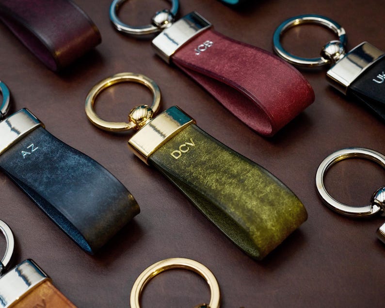 May include: Close-up of several leather keychains in various colors, including burgundy, green, and blue. Each keychain has a metal ring and a leather strap with initials engraved on the strap. The keychains are arranged on a brown surface.