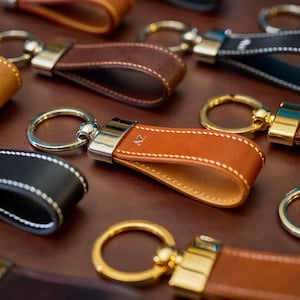 May include: A selection of leather keyrings in shades of brown, black, and tan. Each keyring has a metal ring and a leather loop with contrasting stitching. The keyrings are displayed on a brown surface.