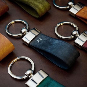 May include: A collection of leather keychains in various colors, including brown, teal, and navy blue. Each keychain features a silver-toned ring and a rectangular metal accent. Some keychains have initials engraved on them.