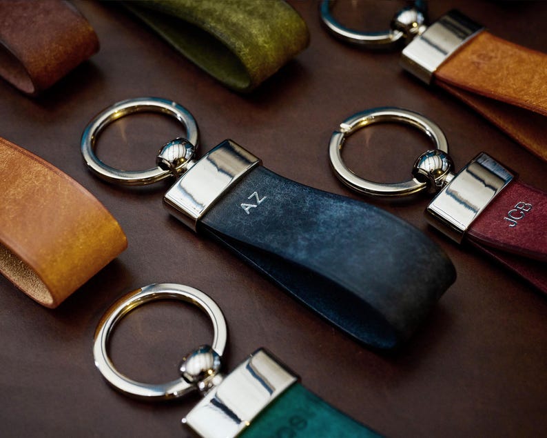 May include: A collection of leather keychains in various colors, including brown, teal, and navy blue. Each keychain features a silver-toned ring and a rectangular metal accent. Some keychains have initials engraved on them.