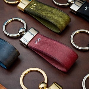 May include: A collection of personalized leather keychains in various colors, including burgundy, green, and black. Each keychain features a silver-toned metal ring and a strap with initials engraved in white. The keychains are arranged on a brown surface.