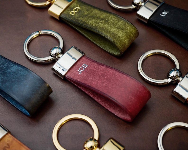 May include: A collection of personalized leather keychains in various colors, including burgundy, green, and black. Each keychain features a silver-toned metal ring and a strap with initials engraved in white. The keychains are arranged on a brown surface.