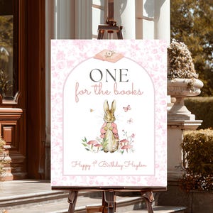 May include: A pink and white sign on an easel with the text "ONE for the books". The sign features a watercolor illustration of a rabbit in a pink coat, mushrooms, and butterflies. The text "Happy 1st Birthday Hayden" is at the bottom.