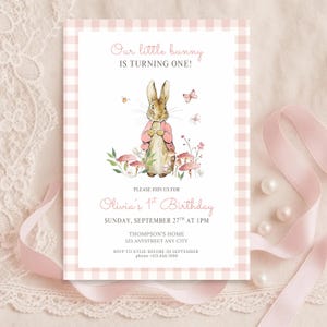 May include: A pink and white gingham patterned invitation card with the text "Our little bunny IS TURNING ONE!". The card features a watercolor illustration of a bunny wearing a pink jacket. The text "Olivia's 1st Birthday" is also on the card.